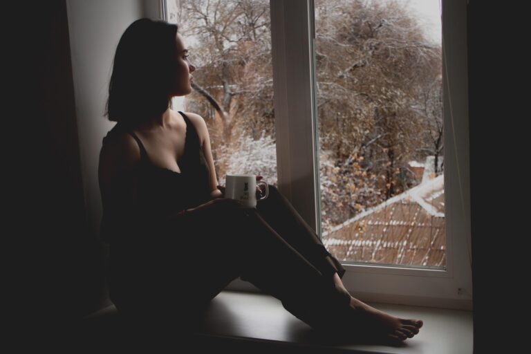 A woman is sitting on a wide windowsill drinking coffee looking out the window.