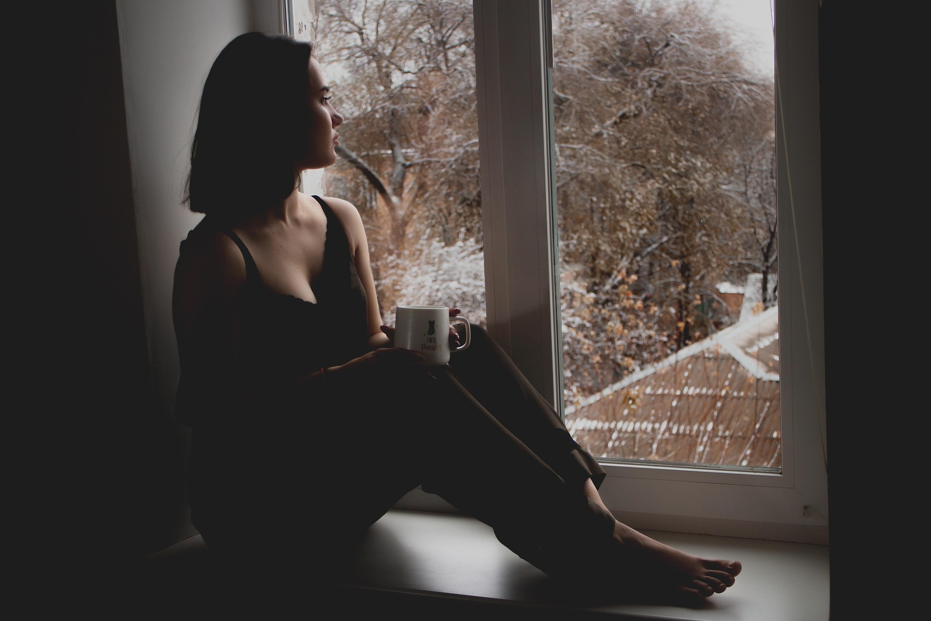 A woman is sitting on a wide windowsill drinking coffee looking out the window.