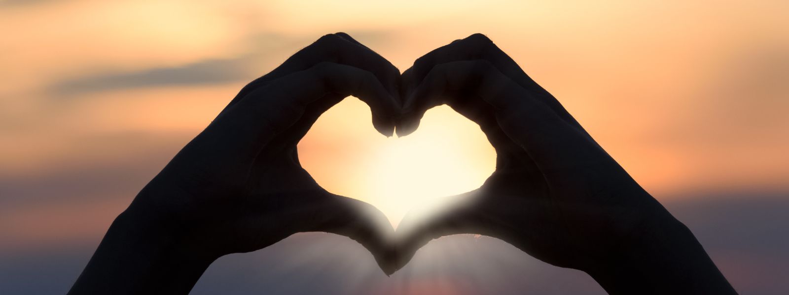 Hands forming a heart around sunlight during a warm sunset, symbolizing hope and healing